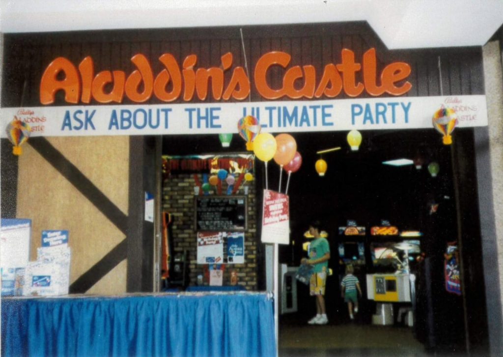 Front entrance to Aladdin's Castle arcade with visible games and a sign that says "Ask about the ultimate party" with patrons in front
