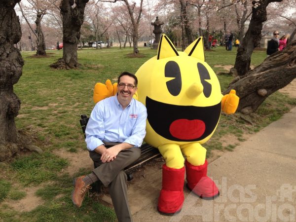 David Bishop wearing a blue Namco shirt, sitting on a park bench with a large Pac-Man mascot