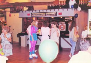 A pink-uniformed employee on roller skates checks on customers in front of the Crossbow Shoot game at Barrel of Fun #13, Commander Ragtime's