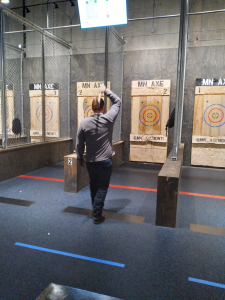 A man in a gray long-sleeve shirt raises an axe overhead, preparing to throw it at a wooden target in an indoor axe-throwing lane marked "MN AXE