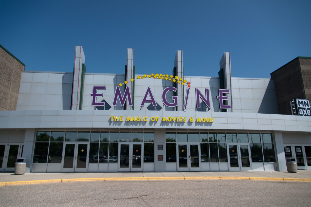 Exterior of the Emagine theater with its purple "EMAGINE" sign and yellow "THE MAGIC OF MOVIES & MORE" tagline under a clear blue sky