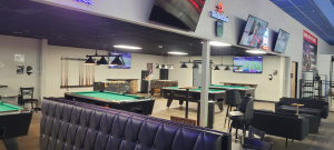 Game room with green-felt pool tables, a foosball table, wall-mounted TVs, and a tufted leather booth in front