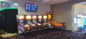 Row of arcade pinball machines, including a Venom-themed table, with a wall TV showing a February 2026 leaderboard